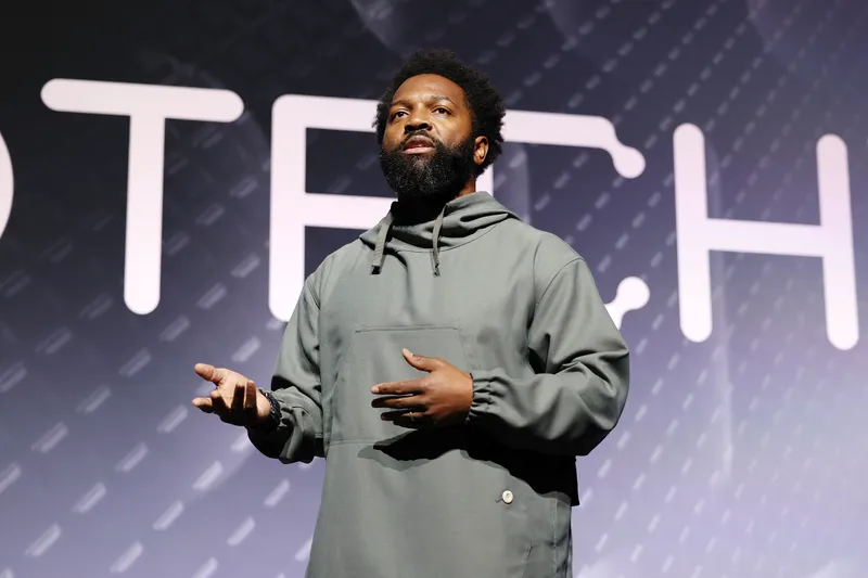 Baratunde Thurston speaking on a conference stage with large tech lettering behind him